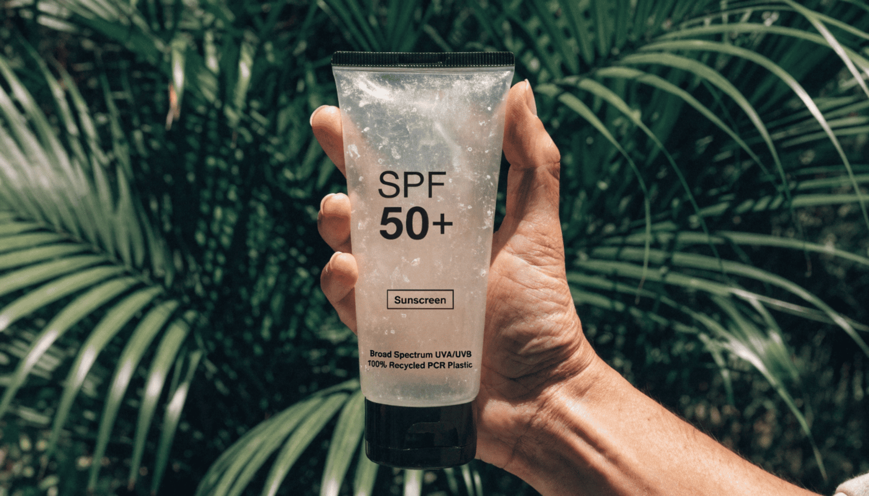 A hand holding a slightly speckled recycled plastic sunscreen tube with a green plant in the background representing sustainability